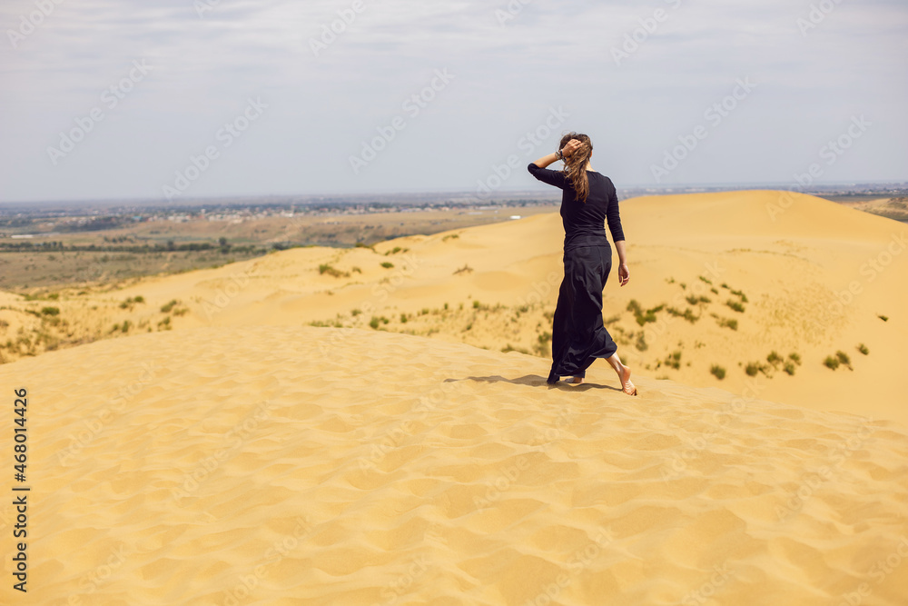 woman in black long dress walks through the desert dunes in summer with her back woman in black long dress walks through the desert dunes in summer with her back