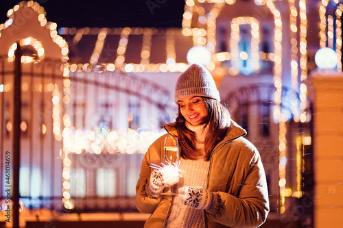 Smiling girl burns sparkler by holiday illumination on new years eve while holds glass of champagne