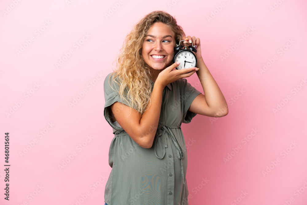 Fototapeta premium Girl with curly hair isolated on pink background pregnant and holding clock