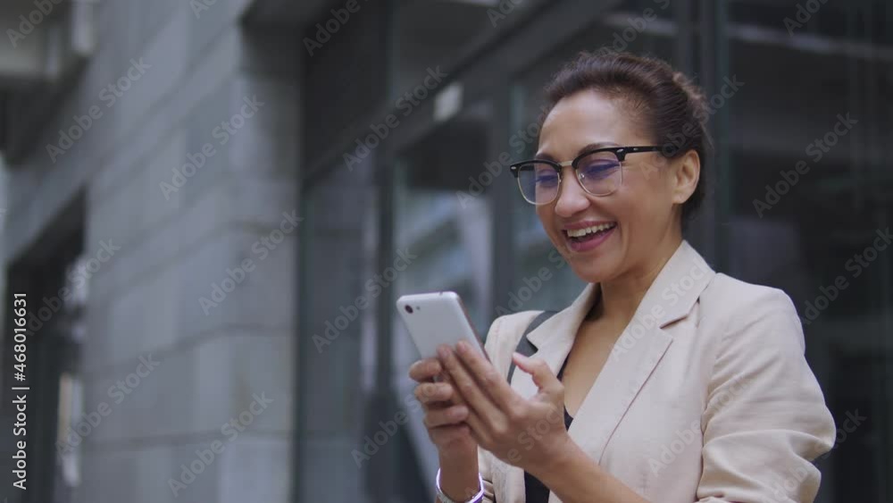 Attractive african american woman in fashionable suit and eyeglasses sincerely smiling standing outdoor with smartphone in hands, reading good news, receiving job promotion and salary increase Attractive african american woman in fashionable suit and eyeglasses sincerely smiling standing outdoor with smartphone in hands, reading good news, receiving job promotion and salary increase