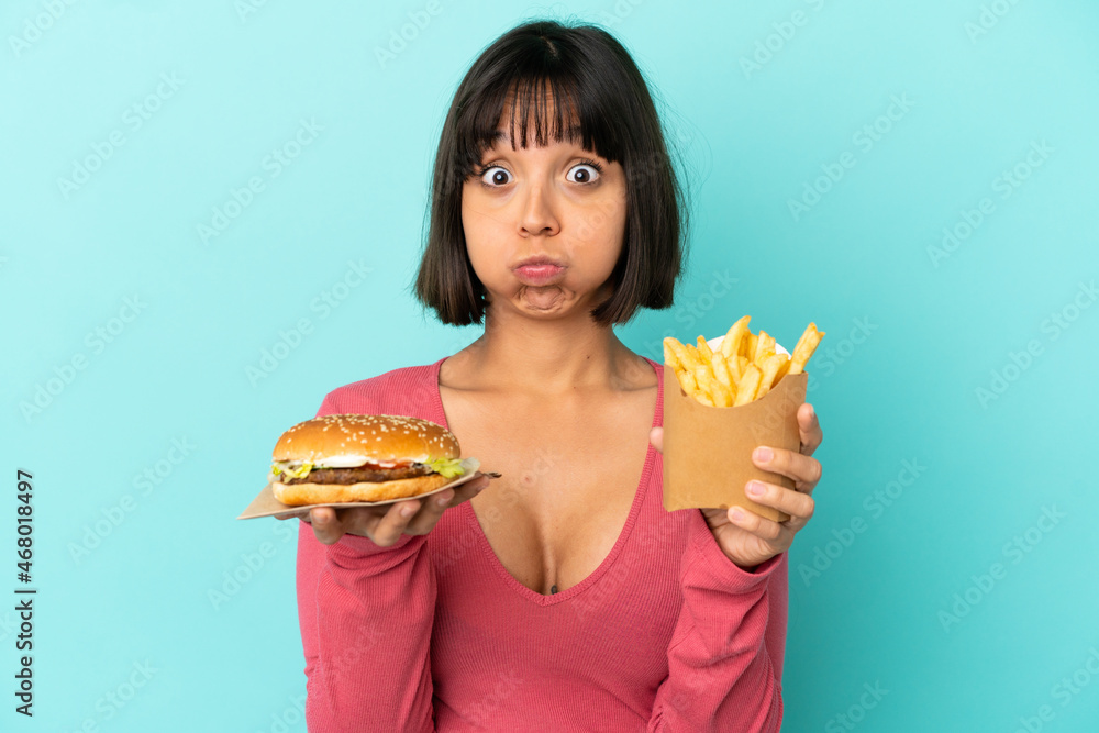 Young brunette woman holding burger and fried chips over isolated blue background Young brunette woman holding burger and fried chips over isolated blue background