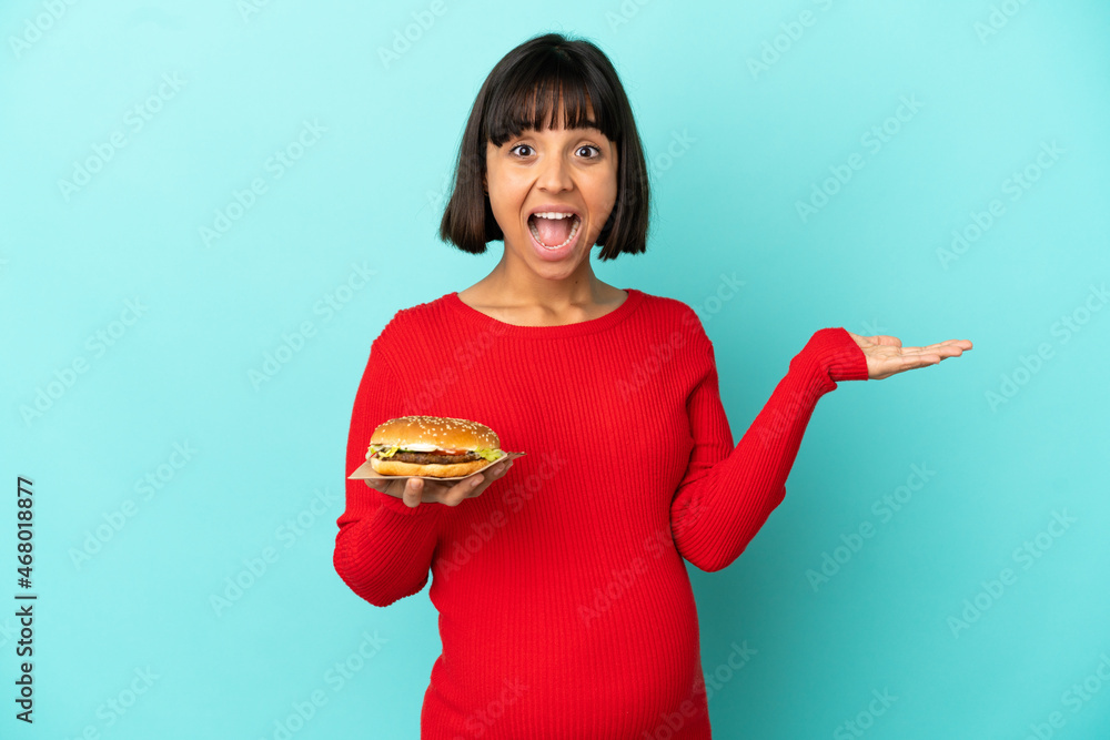 Young pregnant woman holding a burger over isolated background with shocked facial expression Young pregnant woman holding a burger over isolated background with shocked facial expression