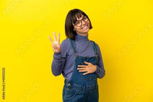 Young pregnant woman over isolated yellow background happy and counting three with fingers