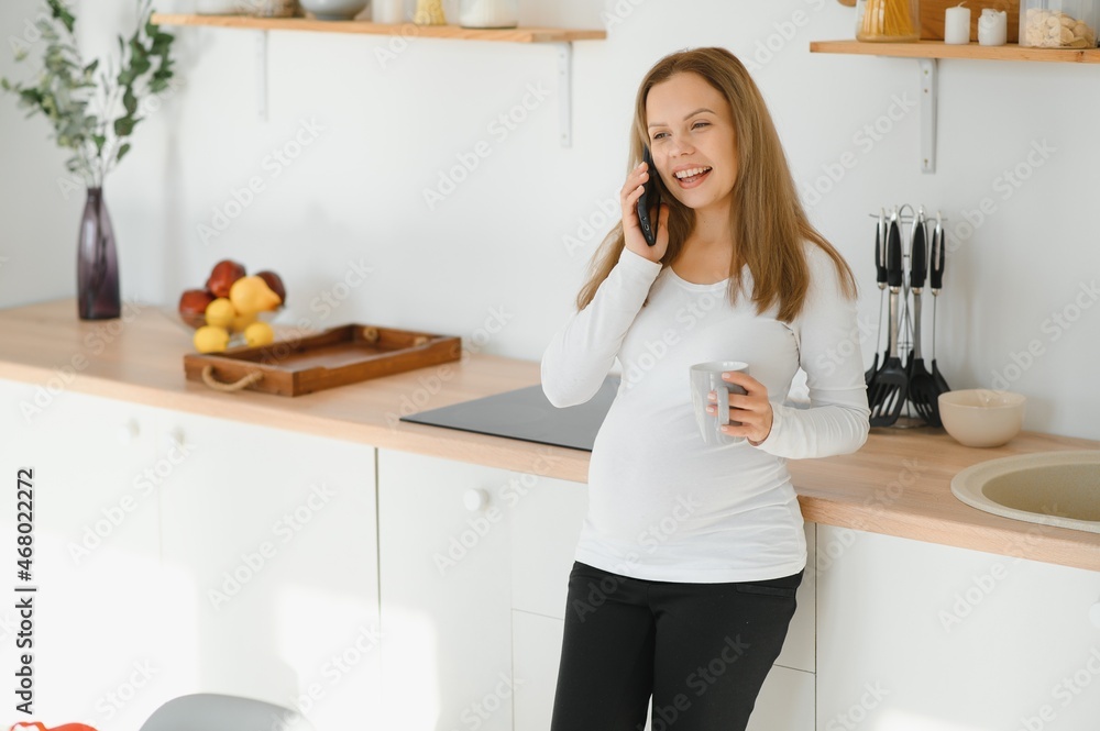 So nice to hear you Beautiful young pregnant woman talking on phone and looking away with smile while standing at kitchen interior