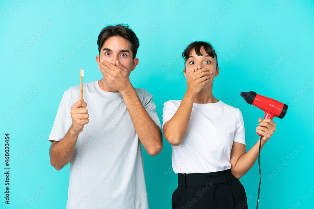 Young mixed race couple holding a hairdryer and toothbrush isolated on blue background covering mouth with hands for saying something inappropriate Young mixed race couple holding a hairdryer and toothbrush isolated on blue background covering mouth with hands for saying something inappropriate