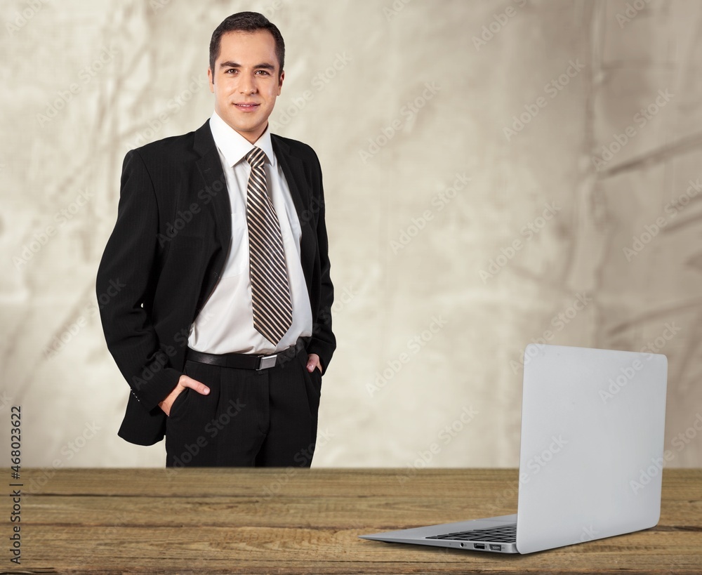 Young businessman in formal suit stands in modern office near desk with a laptop Young businessman in formal suit stands in modern office near desk with a laptop