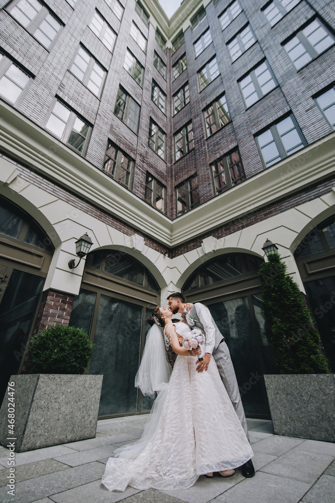 A young bride in a white dress with a long veil and a stylish bearded groom in a gray suit with a bow tie are hugging and kissing against the backdrop of architecture, red brick houses with windows.