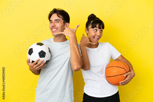 young sport couple playing football and basketball isolated on yellow background making phone gesture. Call me back sign