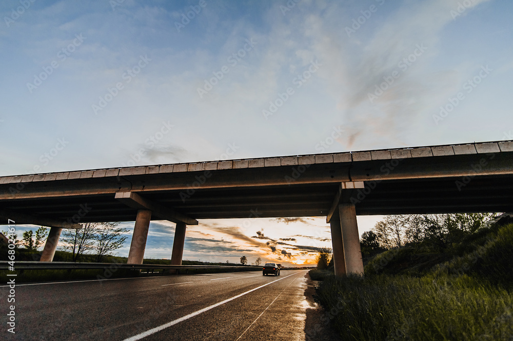 Beautiful nature landscape with a traveling car on a wet asphalt road after rain with a concrete bridge against the backdrop of a sunset and a blue sky with clouds.