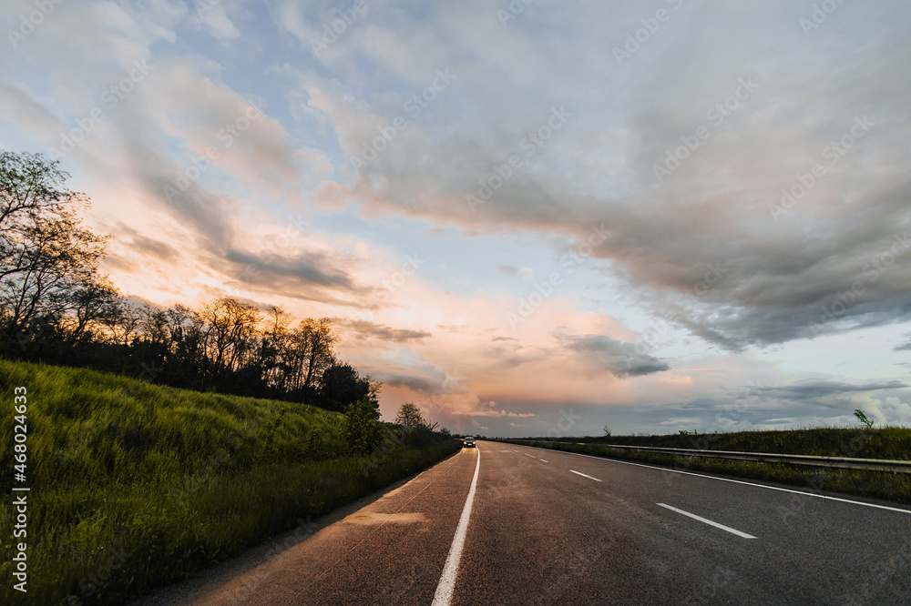 A broken car stands on the side of the road on a wet asphalt road after rain against the backdrop of a sunset and a blue sky with clouds. Beautiful landscape. Evening photography.
