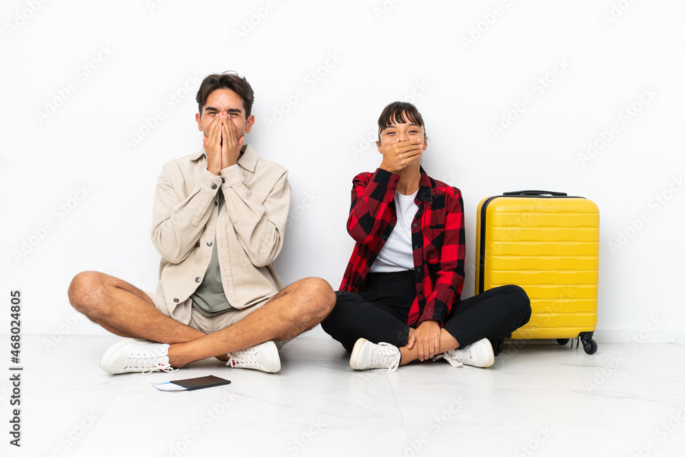 Obraz premium Young mixed race travelers couple sitting on the floor isolated on white background smiling a lot while covering mouth