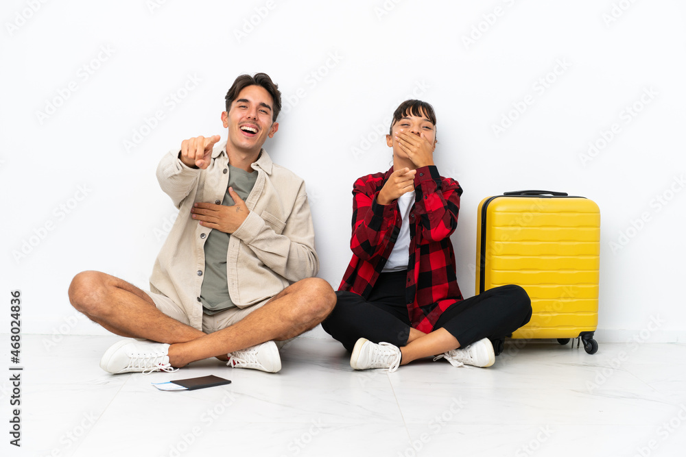 Young mixed race travelers couple sitting on the floor isolated on white background pointing with finger at someone and laughing a lot Young mixed race travelers couple sitting on the floor isolated on white background pointing with finger at someone and laughing a lot