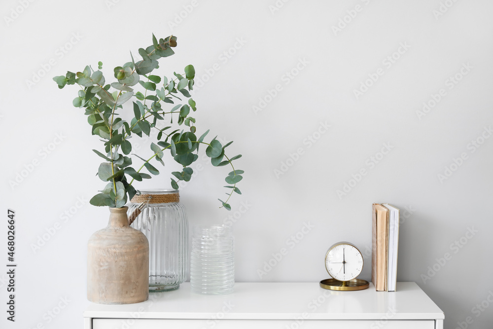 Vases with eucalyptus branches, alarm clock and books on shelf near light wall