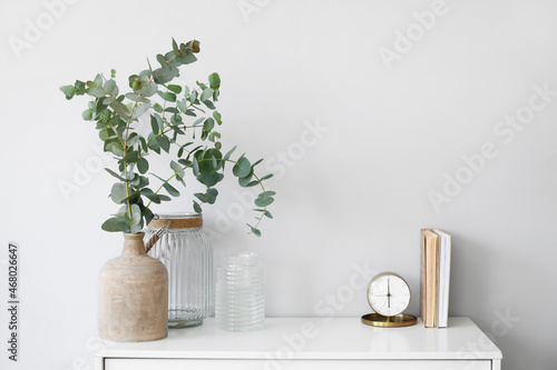 Vases with eucalyptus branches, alarm clock and books on shelf near light wall © Pixel-Shot
