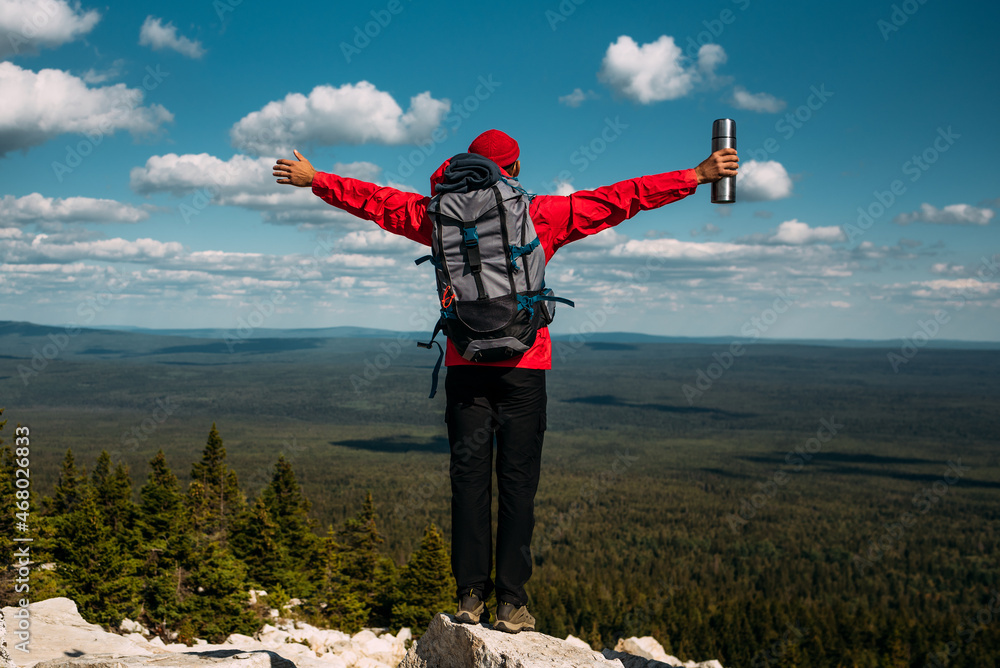 A tourist traveler with his hands raised stands on the top of the mountain. A male traveler with a backpack in the mountains, rear view. A trip to the mountains with a backpack. Copy space