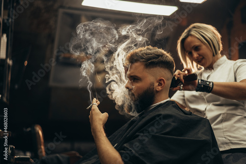 Young bearded man getting haircut by hairdresser and smoking a cigarette
