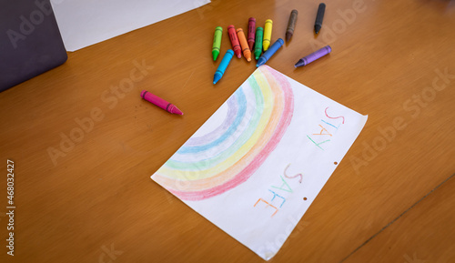 A rainbow painting with a text Stay Safe lying on a table with colourful crayons lying around in a m