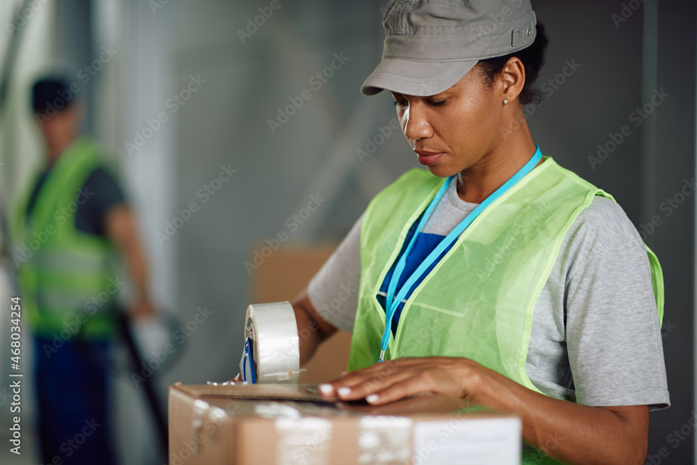 African American warehouse worker uses tape dispenser while preparing boxes for shipment and working at distribution compartment. African American warehouse worker uses tape dispenser while preparing boxes for shipment and working at distribution compartment.