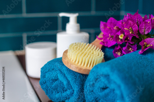 Two rolls of aquamarine terry towels, a beautiful flower and a wooden bath brush and other hygiene items out of focus are on the shelf in the bathroom against the backdrop of tiles. Spa theme.