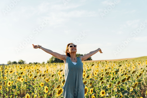 Middle-aged young woman wearing sunglasses, breathing fresh air with open arms in sunflower field