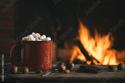 Cocoa with marshmallows and chocolate in a red mug on a wooden table near a burning fireplace
