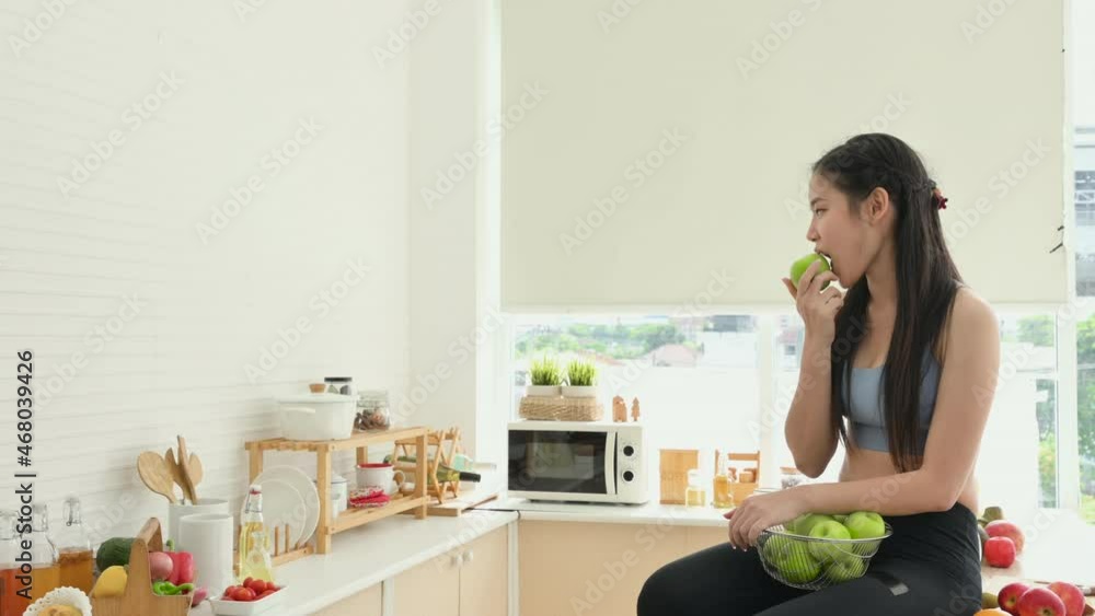 Happy Asian healthy woman in sportswear holding basket and eating green apple in kitchen at apartment. Beautiful sports girl sit smile on cook table and enjoy eat fruit for health after exercise.
