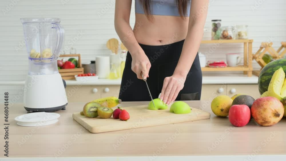 Healthy young woman in sportswear cutting green apple on chopping board and putting into blender while preparing smoothie by kitchen table. Sports girl preparing fruit and ingredients for smoothie. Healthy young woman in sportswear cutting green apple on chopping board and putting into blender while preparing smoothie by kitchen table. Sports girl preparing fruit and ingredients for smoothie.