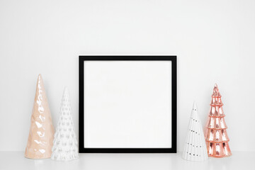Christmas mock up with black square frame with white and rose tree decor on a white shelf against a white wall. Copy space.