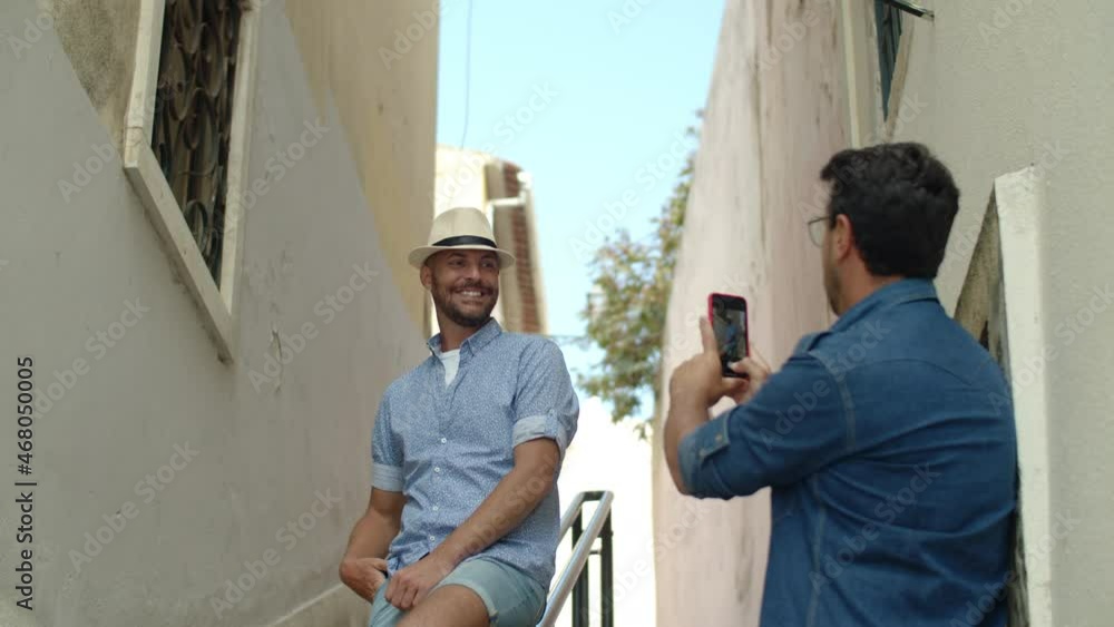 Medium shot of handsome gay taking picture of boyfriend outdoor. Smiling homosexual man standing in narrow street of old Spain village on summer day and posing for camera. LGBT, photoshoot concept Medium shot of handsome gay taking picture of boyfriend outdoor. Smiling homosexual man standing in narrow street of old Spain village on summer day and posing for camera. LGBT, photoshoot concept
