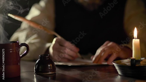 A scene from the 18th century of a man writing with a quill pen with a candle and a hot beverage sitting on his desk.