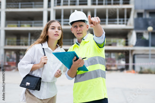 Professional builder man talking with woman client while standing at construction site, pointing to something