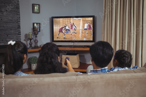 Rear view of family sitting at home together watching basketball match on tv
