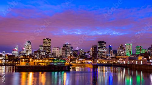 Photo de la ville de Montréal prise au début de la nuit. Le ciel est d'un bleu intense, les nuages rosés et les lumières de la ville se reflètent sur le fleuve Saint-Laurent.