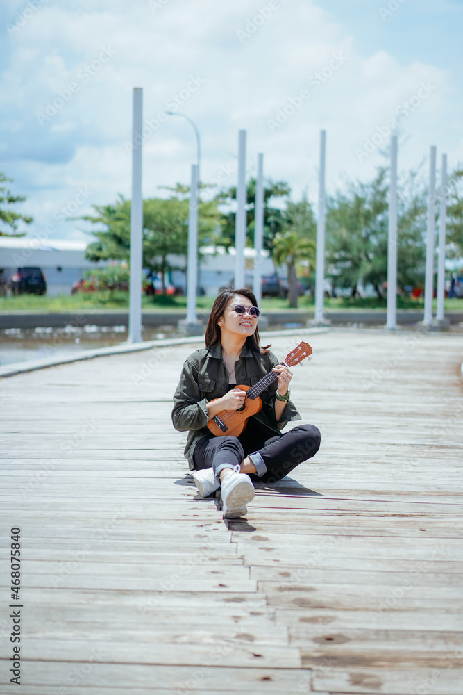 Fototapeta premium Playing Ukulele of Young Beautiful Asian Woman Wearing Jacket And Black Jeans Posing Outdoors