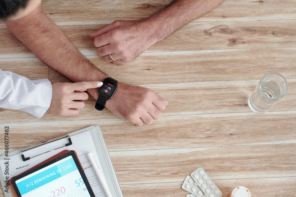 Doctor showing aged patient how to check heart rate on his smartwatch, view from the top Doctor showing aged patient how to check heart rate on his smartwatch, view from the top