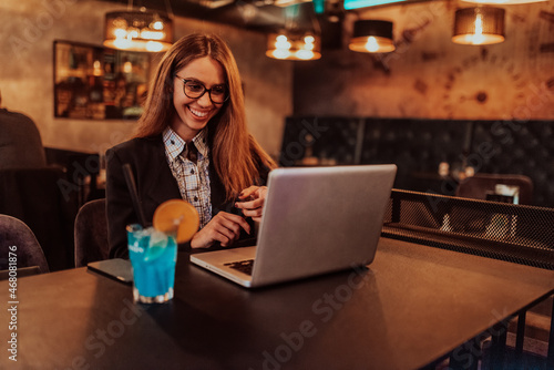 Business woman in glasses sitting in a modern cafe and working on her laptop while drinking a cocktail. Selective focus