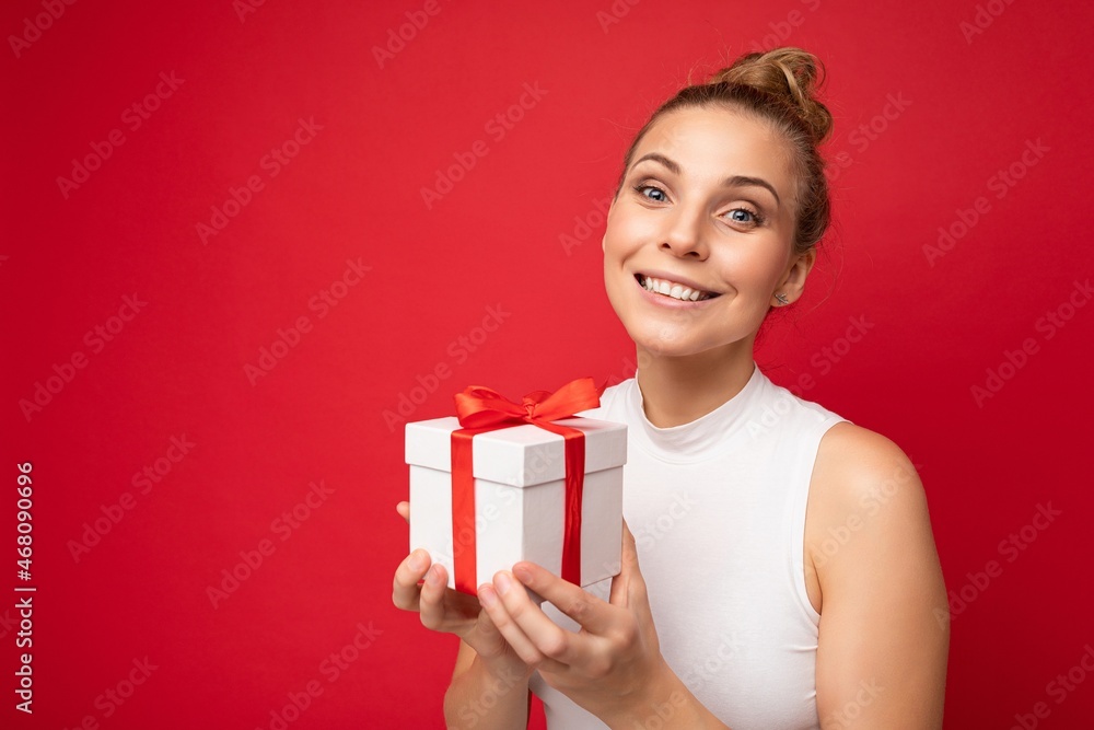 Photo shot of beautiful attractive cute positive happu smiling young blonde woman isolated over red background wall with copy space for text wearing casual white top holding gift box with red ribbon Photo shot of beautiful attractive cute positive happu smiling young blonde woman isolated over red background wall with copy space for text wearing casual white top holding gift box with red ribbon