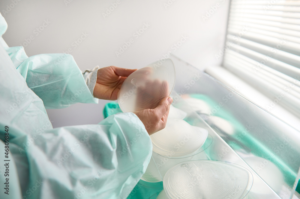 Breast prothesis in surgeon hands on background of medical samples of breast silicone implants of various sizes for breast augmentation aesthetic plastic surgery after resection due to cancer disease Breast prothesis in surgeon hands on background of medical samples of breast silicone implants of various sizes for breast augmentation aesthetic plastic surgery after resection due to cancer disease