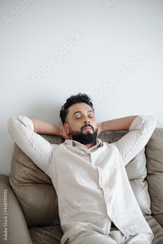 Tired man in white shirt closing eyes and leaning back on comfy sofa