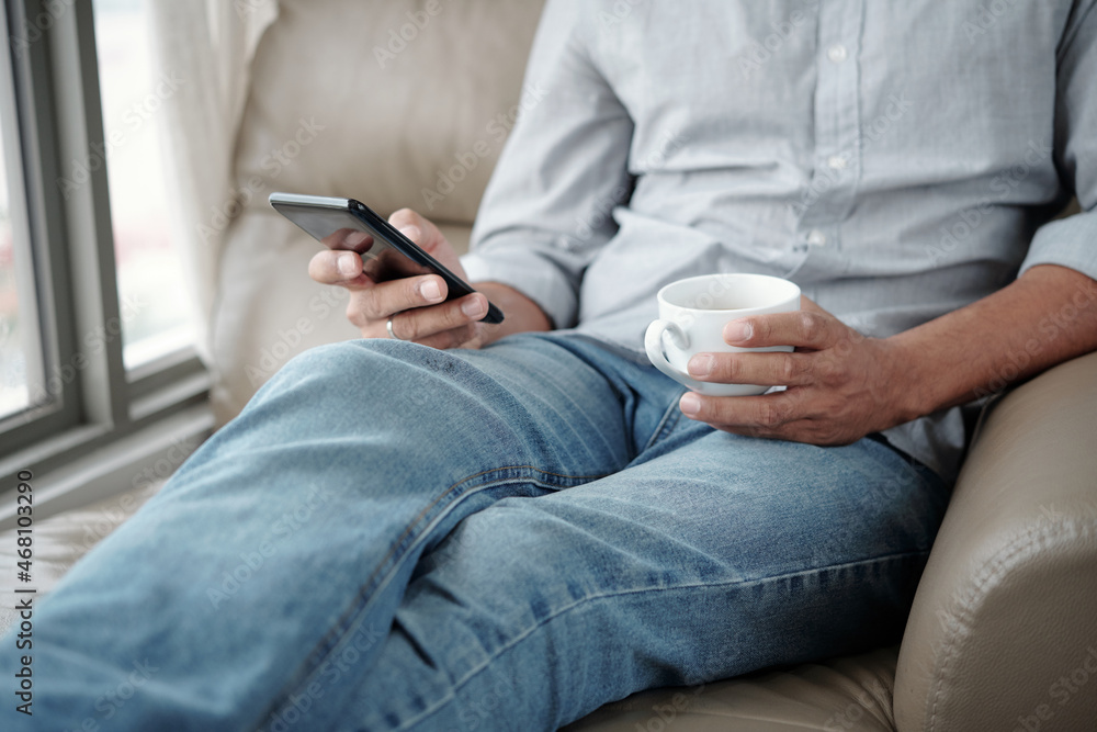 Cropped image of man sitting in armchair, drinking cup of coffee and checking social media on smartphone Cropped image of man sitting in armchair, drinking cup of coffee and checking social media on smartphone