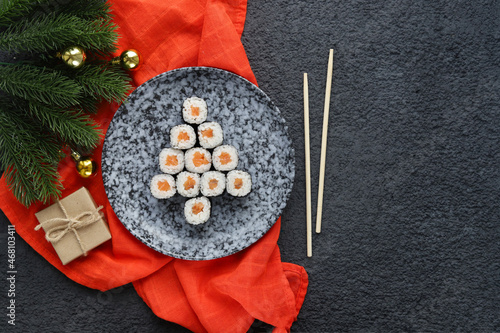 Christmas tree made of sushi on gray plate on dark background among branches of Christmas tree. A creative idea for Japanese restaurant. The concept of Christmas and New Year dishes.Copy Space