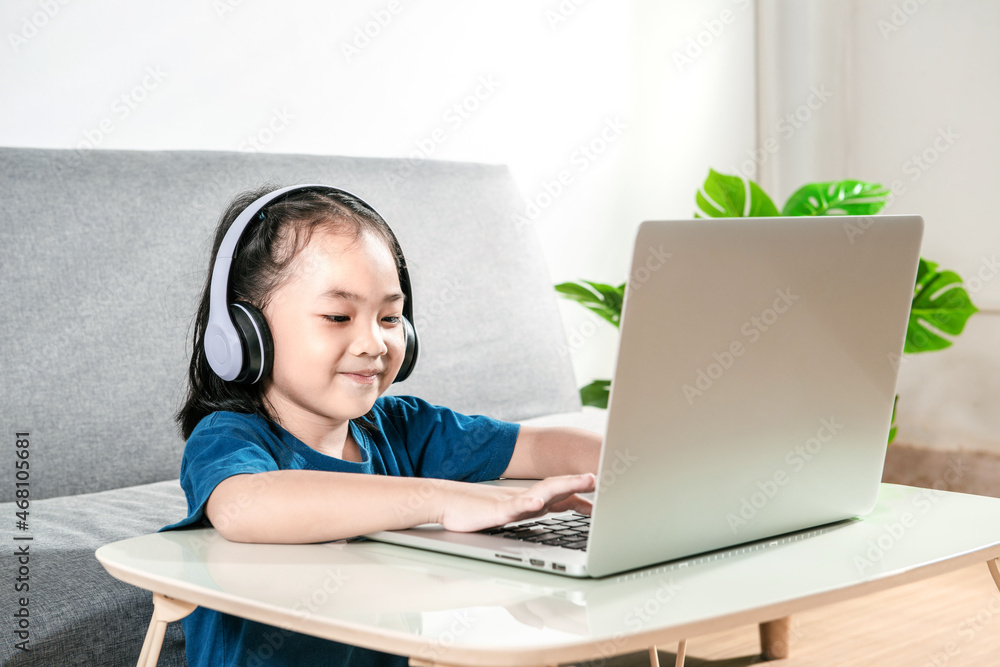 Cheerful Little kid girl studying online class with laptop and headphones at home. A New normal, Covid-19 coronavirus. Social Distance learning education technology gaming and homeschooling concept Cheerful Little kid girl studying online class with laptop and headphones at home. A New normal, Covid-19 coronavirus. Social Distance learning education technology gaming and homeschooling concept