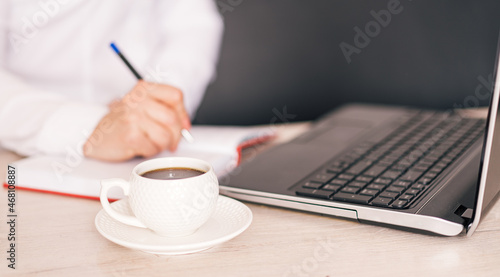 Woman writes with pen in notebook in front of laptop and cup of coffee.