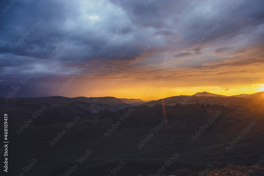 Atmospheric landscape with silhouettes of mountains with trees on background of dawn sky with vivid orange sunlight and sun rays. Colorful nature scenery with sunset or sunrise of illuminating color.