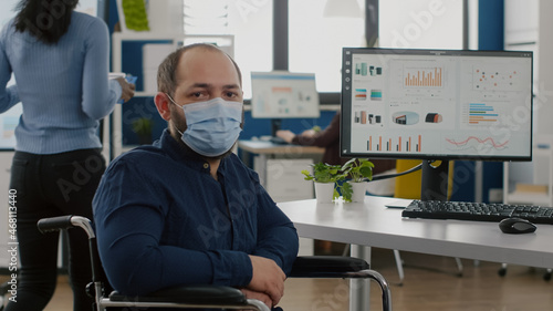 Employee with disabilities, invalid, handicapped paralized with face mask against coronavirus looking at camera sitting immobilized in wheelchair in business office room, working at financial project