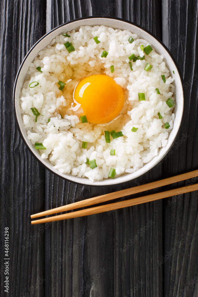 Tamago kake gohan is a popular Japanese breakfast food consisting of cooked Japanese rice topped with raw egg close up in the bowl on the table. Vertical top view from above