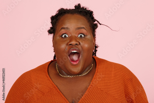 Shocked young black plus size body positive woman opens mouth in amazement wide-eyed standing on light pink background in studio closeup