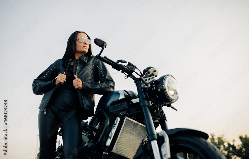Young woman stands near black motorbike against sky background. Young woman stands near black motorbike against sky background.