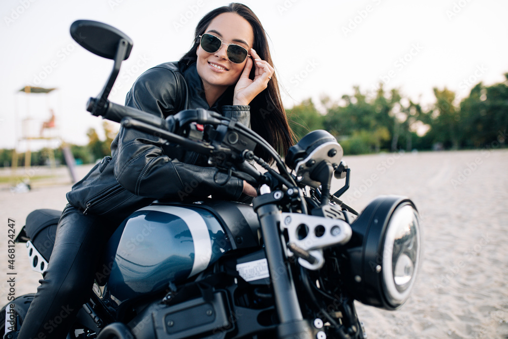 Obraz premium Young woman sits on black motorbike among sandy beach.