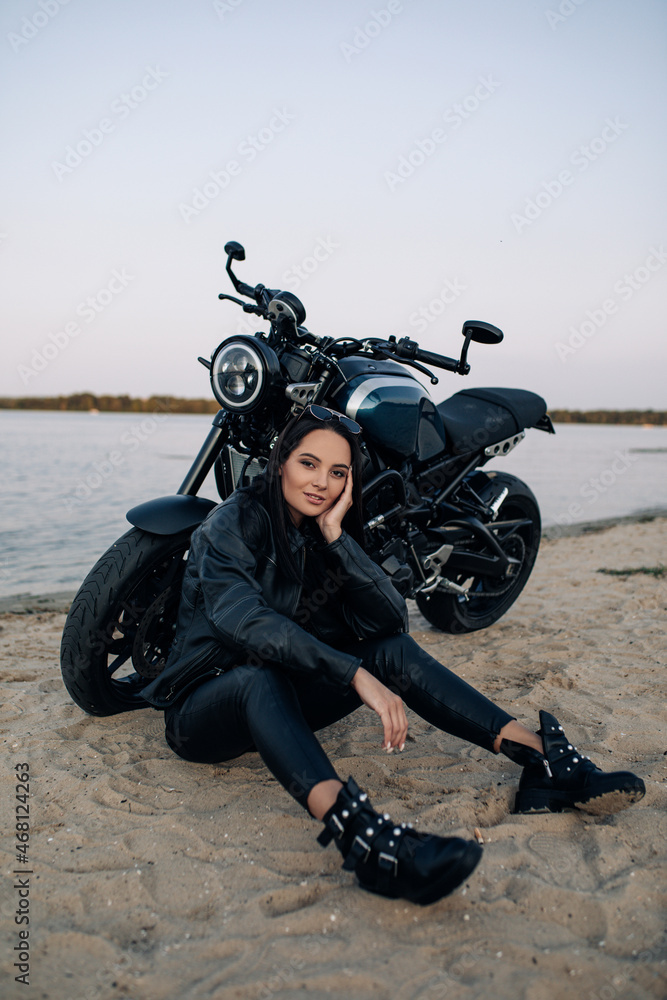 Obraz premium Young woman sits near black motorbike on beach near river.